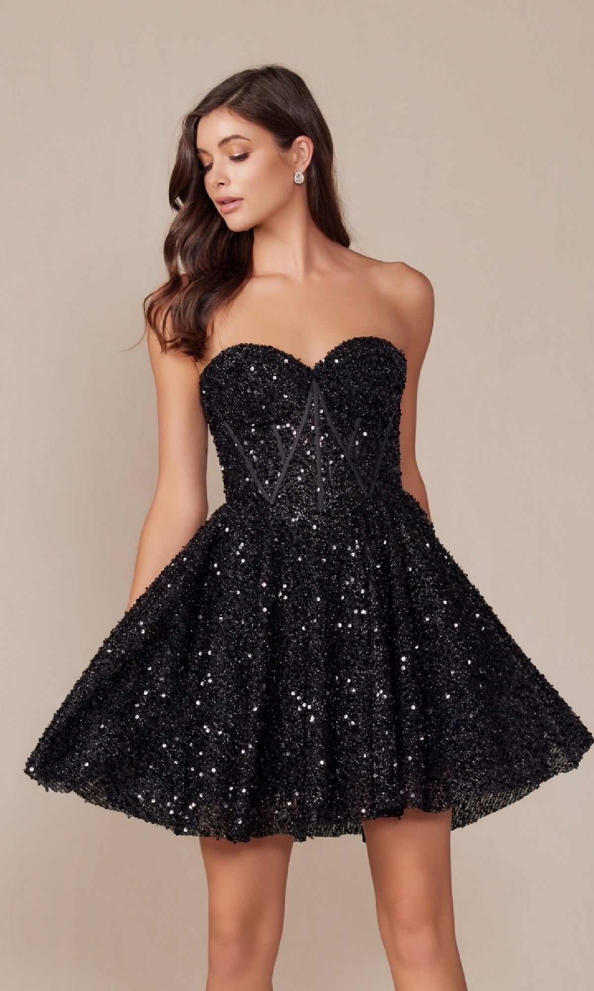 A woman stands against a beige background in the Nox Anabel A843 Sequin Corset Homecoming Dress—strapless, black, with a sequin bodice and flared skirt.